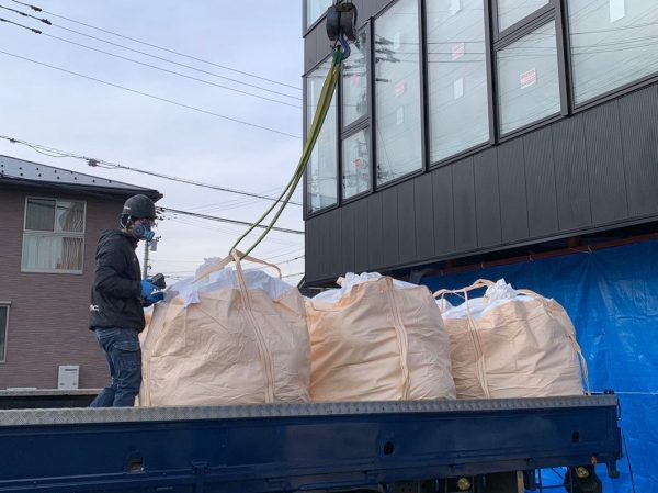 富山のアスベスト解体現場で、株式会社エイキの作業員が廃材の入ったフレコンバックを紺色のトラックの荷台に乗せている。